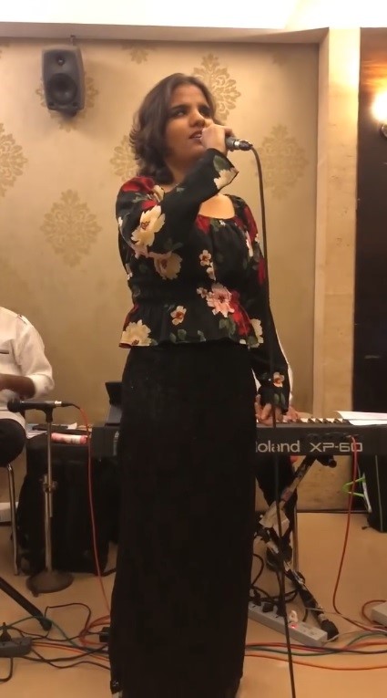 Neha, wearing black floral top and black bottom, holds a black microphone and give live performance. In the background, instrument players are there and play music using the instruments in some function hall.