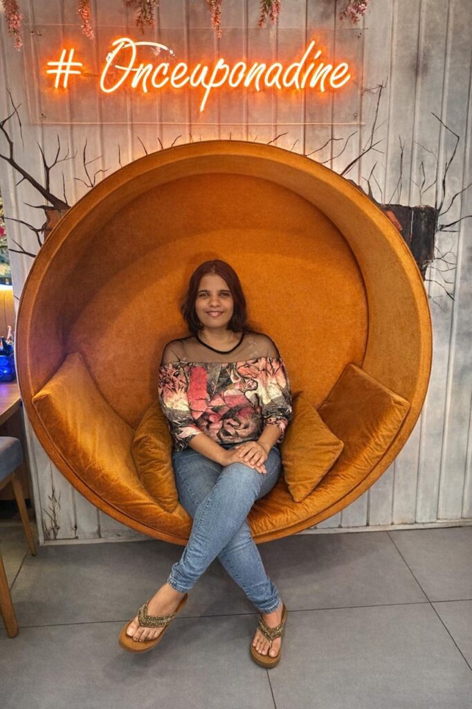 Neha Gianani sits cross-legged on a round orange chair with two cushions, under a neon sign reading "#Onceuponadine" mounted on a wooden wall.